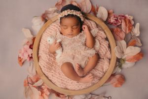 Newborn baby sleeping peacefully in wooden bowl surrounded by pink and peach flowers on soft knit blanket.