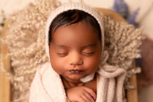 Sleeping newborn in white knit bonnet nestled in cream colored fabric.