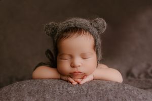 Sleeping newborn wearing knit bear bonnet resting on grey fabric.