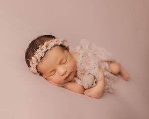 Peaceful newborn photography of a sleeping baby wearing a floral crown and lace outfit on pink background.