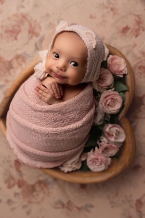 Smiling baby wrapped in pink swaddle posed in basket with roses.
