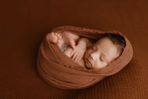 Newborn baby swaddled in soft brown fabric sleeping peacefully on textured brown backdrop.