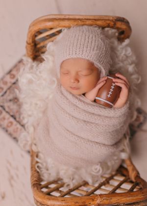 Sleeping newborn wrapped in knit blanket holding miniature football.