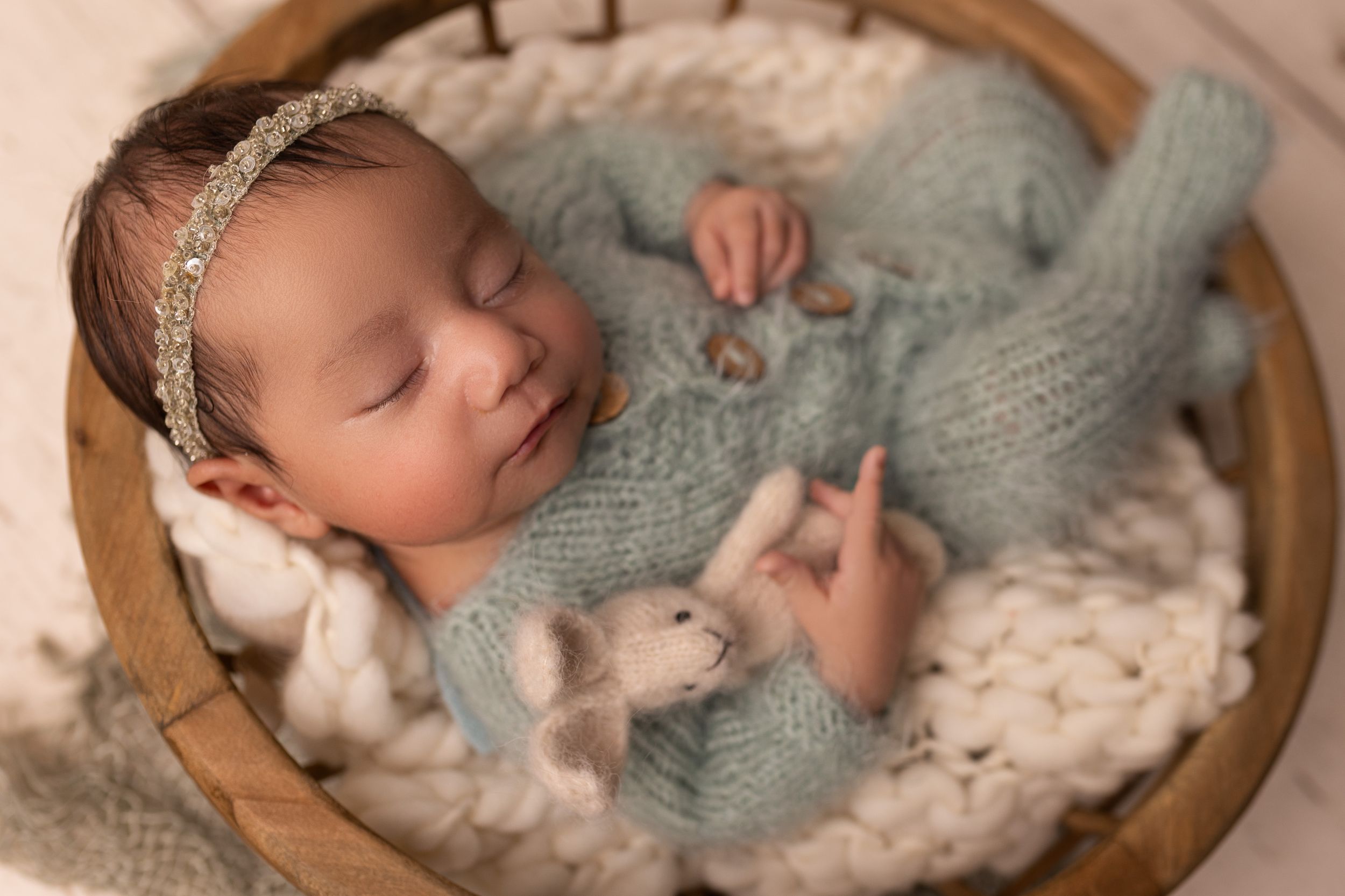 Sleeping newborn in soft blue knit outfit with delicate headband in wooden bowl.