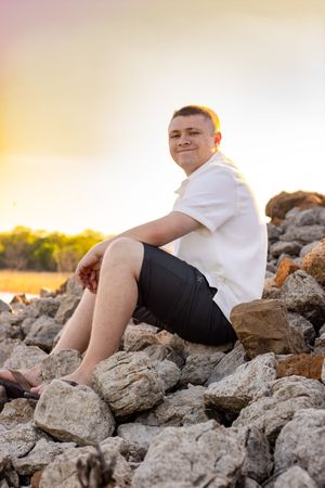 A person in a white shirt and black shorts sits on rocky terrain at sunset.