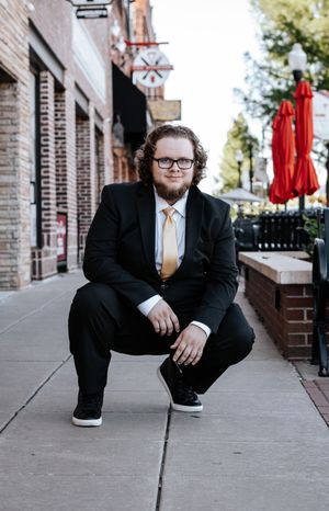 Professional portrait of a person in a black suit and yellow tie crouching on an urban sidewalk near brick buildings.