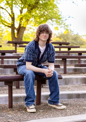 Person in striped shirt and jeans sits casually on wooden park bench with autumn trees in background.