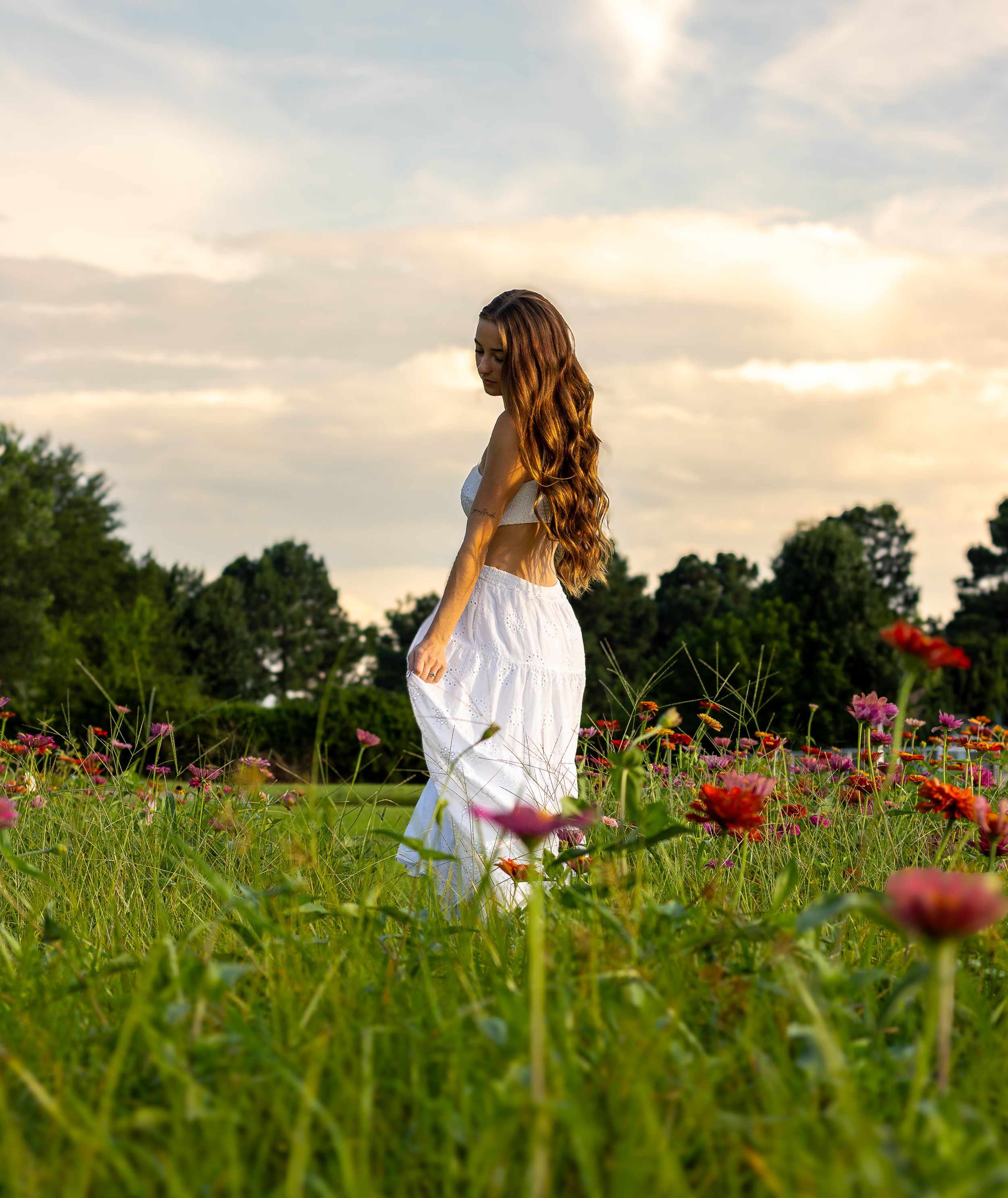 A woman in a flowing white dress stands in a wildflower meadow with red poppies during golden sunset light.