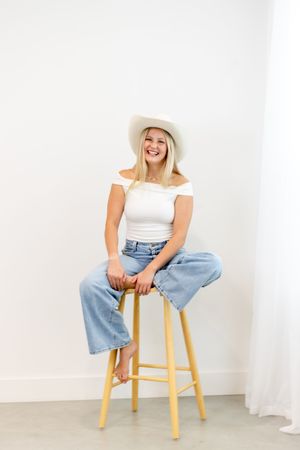 Woman in white top and jeans sits casually on wooden stool against white backdrop, creating relaxed lifestyle portraits.