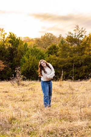 A person wearing a white sweater and jeans poses playfully in a golden field during sunset.