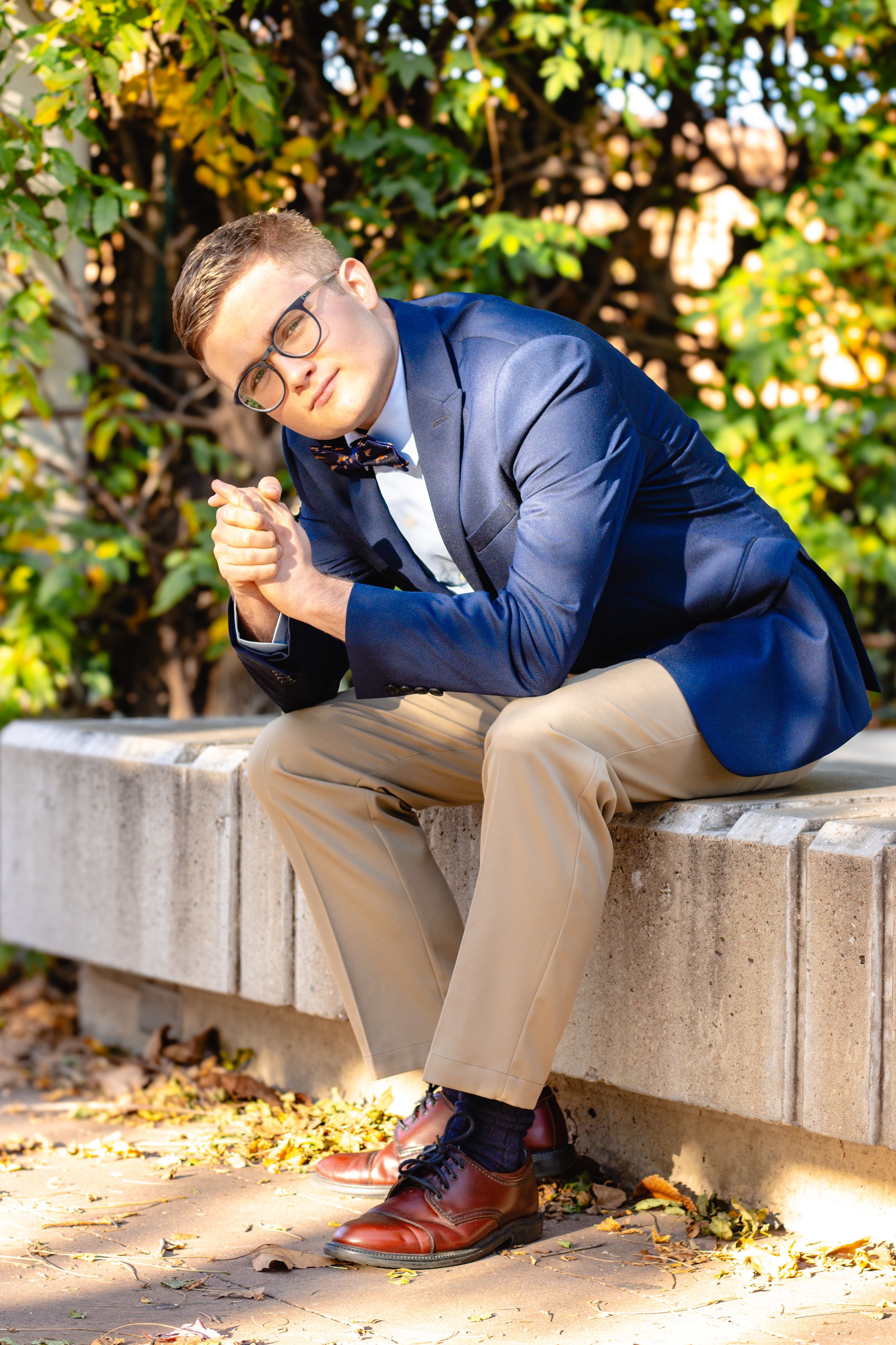 Professional in a navy suit and khakis sits casually on concrete steps in an outdoor setting with autumn leaves.