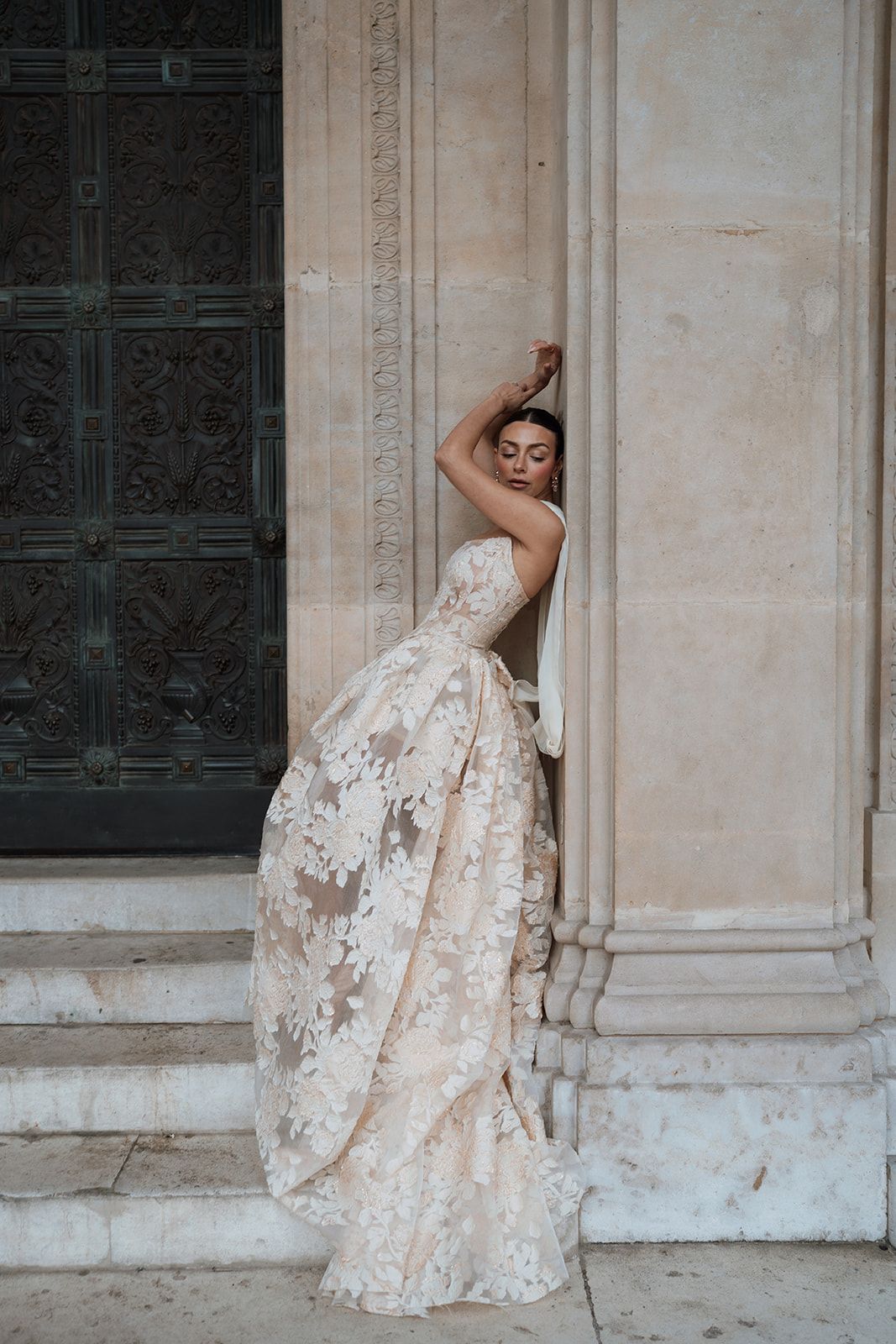 A person in a white wedding dress poses on a balcony with the illuminated Eiffel Tower glowing against the night sky.