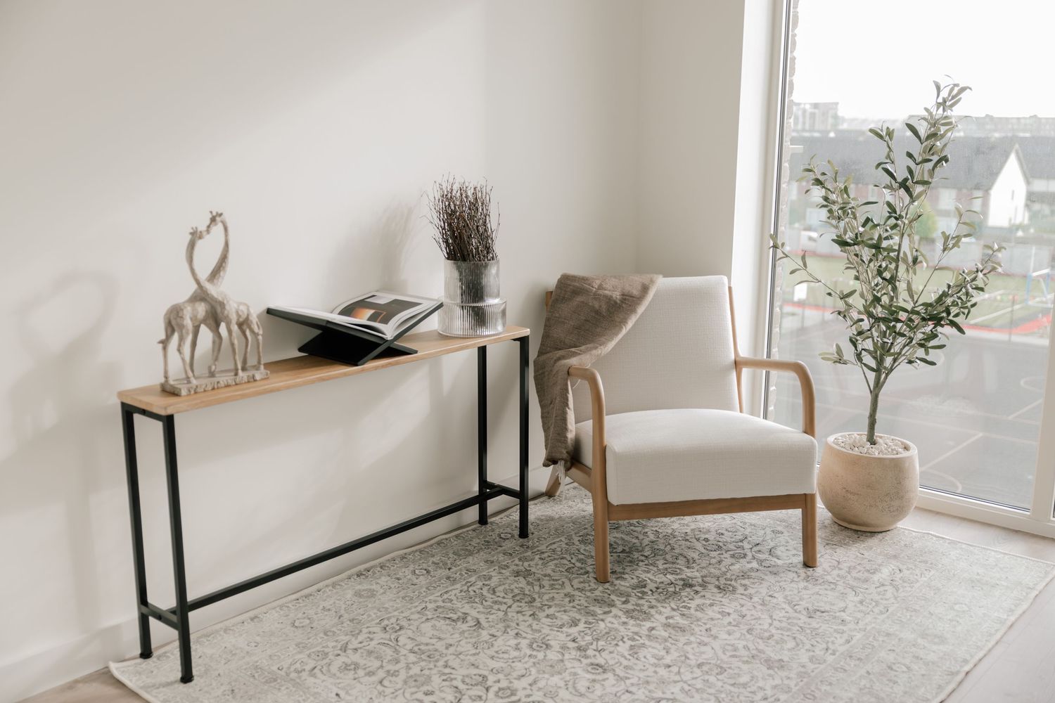 Modern minimalist entryway featuring a slim console table, wooden armchair, and potted plant against white walls.