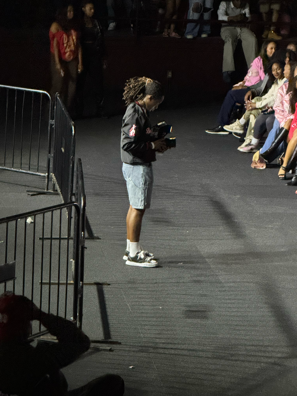 Mills standing on dark stage floor holding camera, with audience seated behind metal barrier.