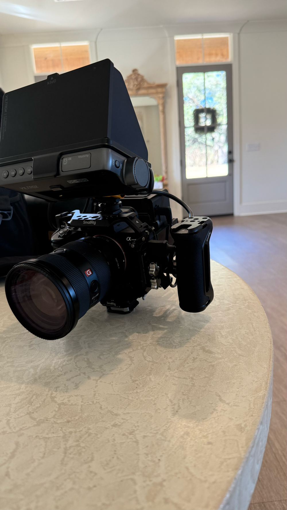 Professional camera rig with viewfinder and lens on a table inside a bright home entryway with a wreath on the door.