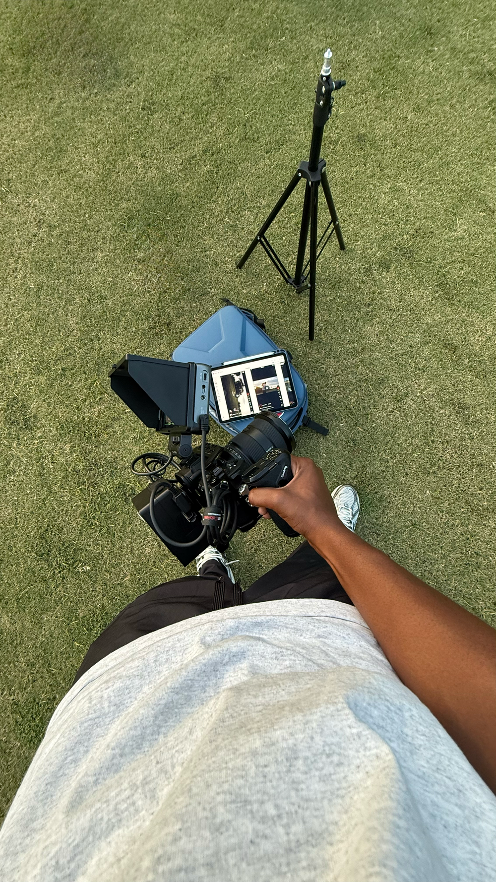 Photographer standing on grass holding a DSLR camera with a tablet monitor and light stand setup outdoors.