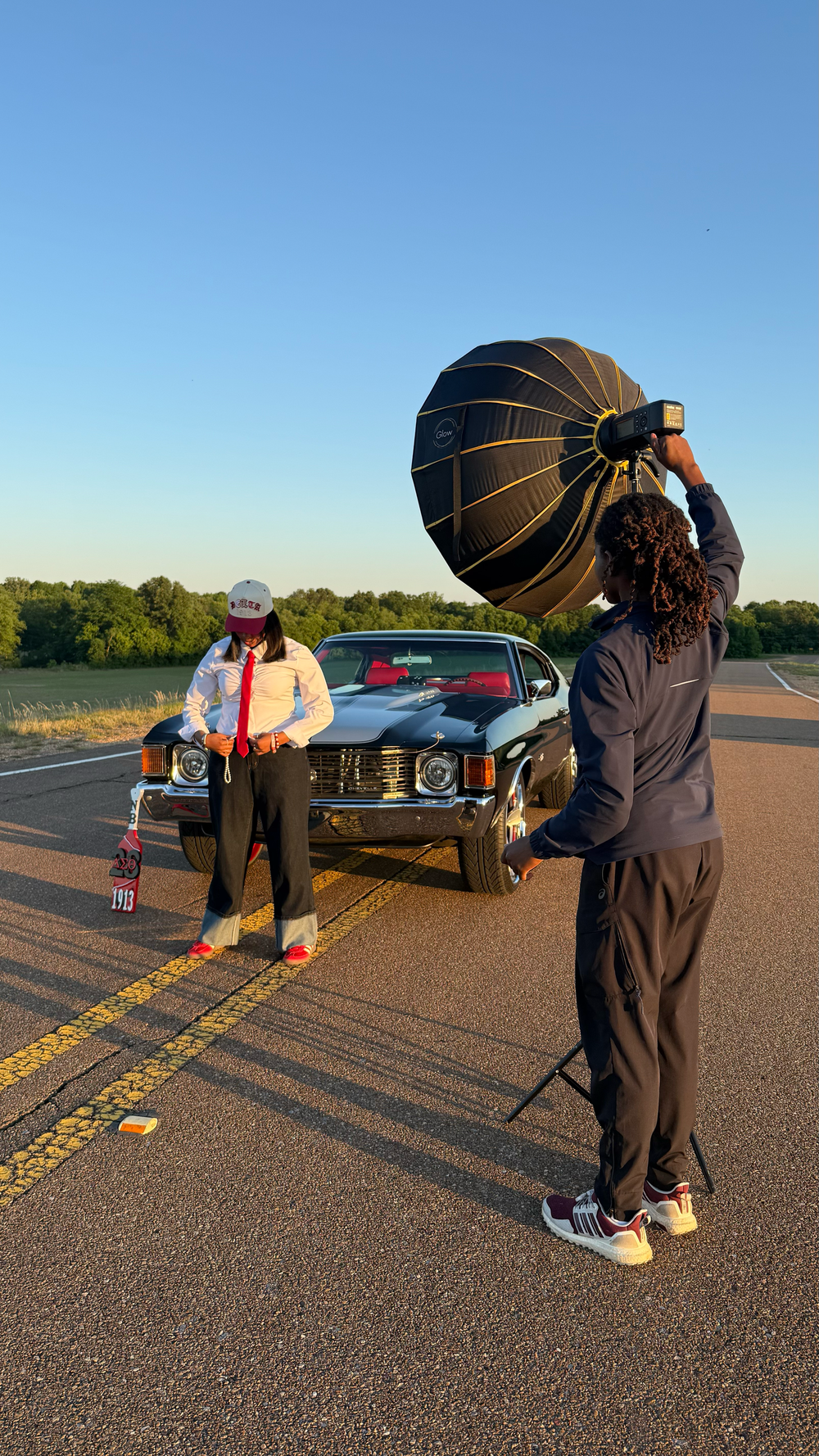 Mills using large softbox light to photograph model posing by classic black car at sunset.