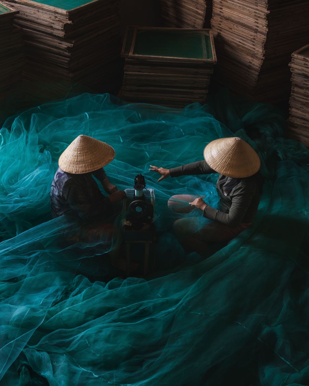 Two workers in conical hats mending fishing nets amid turquoise fabric in Vietnam. Photograph by Chris Ha.