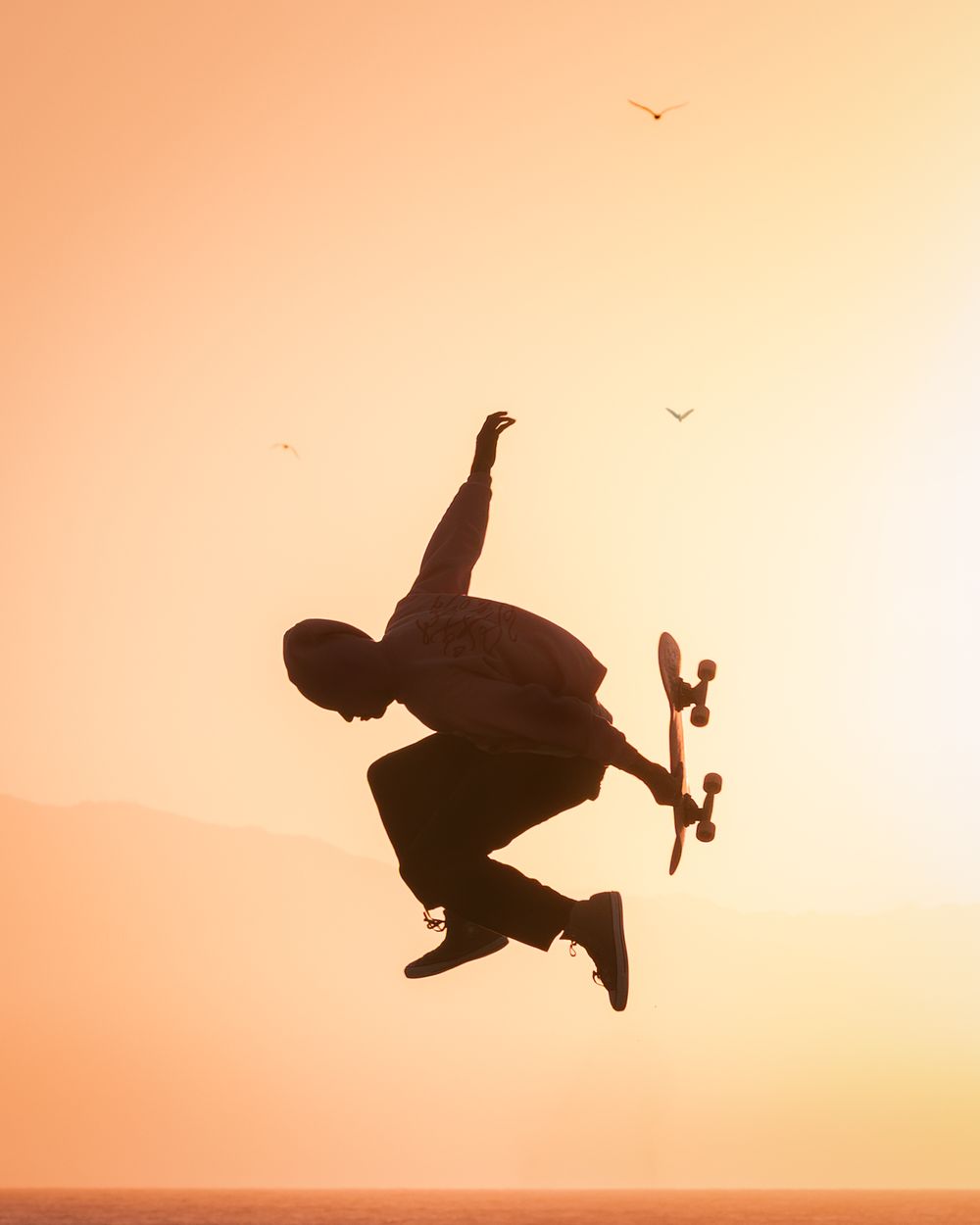 Silhouette of skateboarder performing a jump trick against an orange sunset sky with birds. Photograph by Chris Ha.