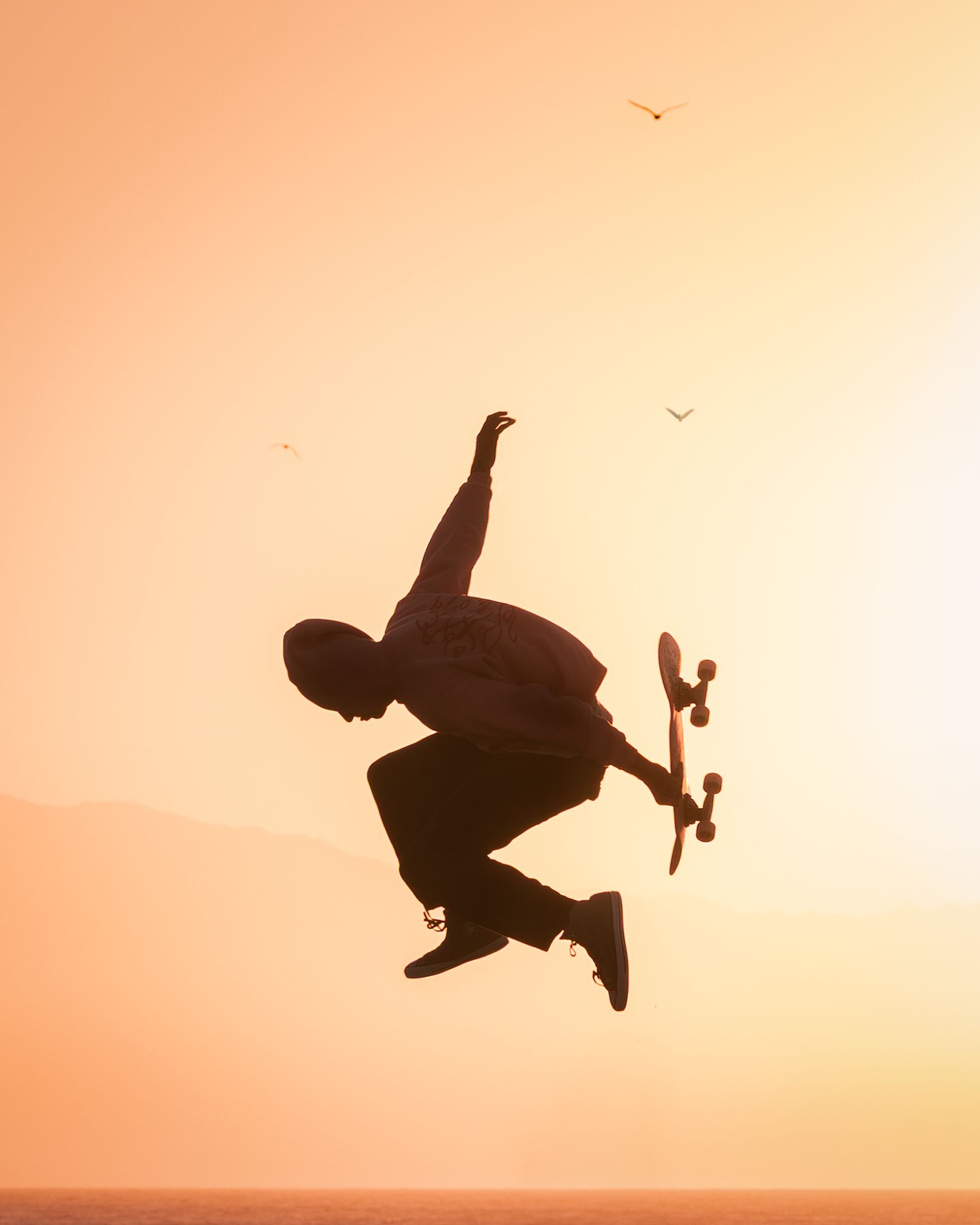 Silhouette of skateboarder performing a jump trick against an orange sunset sky with birds. Photograph by Chris Ha.