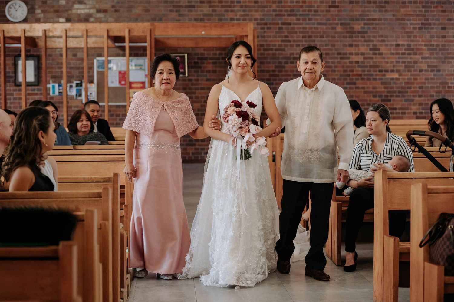 Wedding ceremony in a church with a person in a white wedding gown walking down the aisle with two others in formal attire.