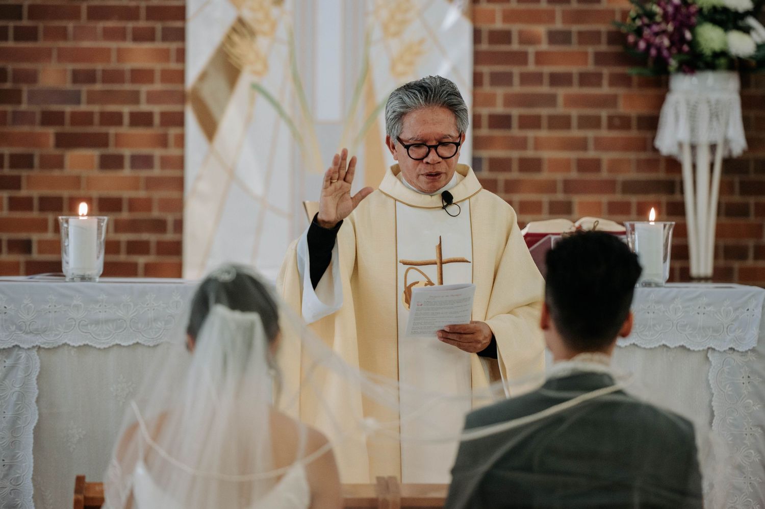 A clergy member in cream vestments leads a wedding ceremony at the altar of a brick church.