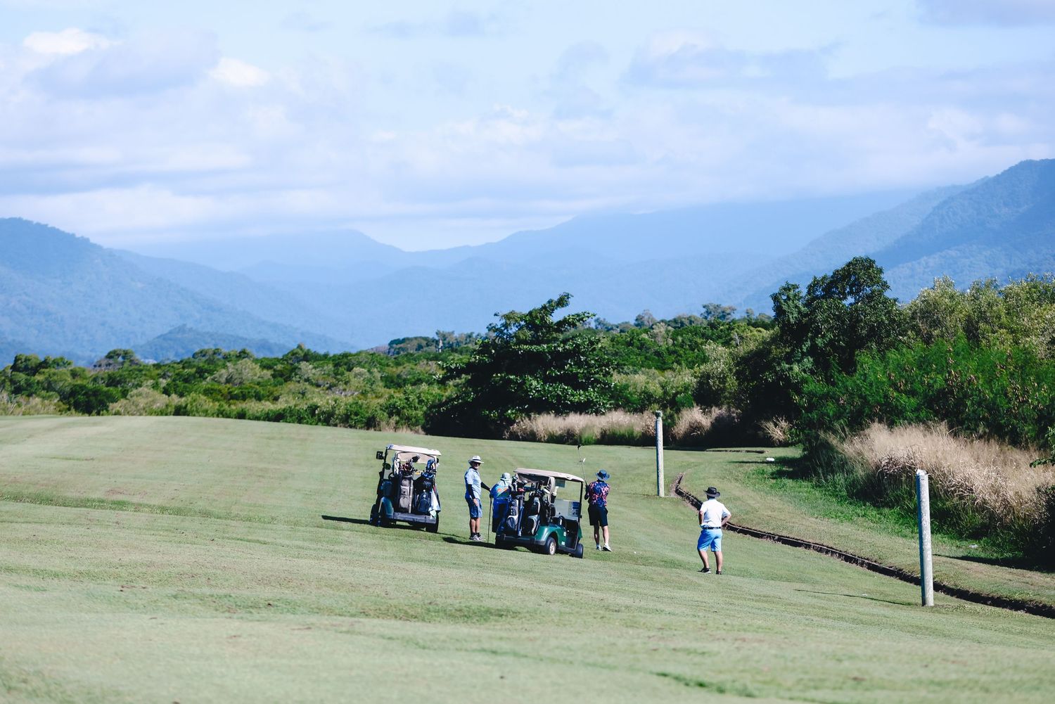 Golfers and spectators gather on a scenic hilltop golf course with mountains and ocean views in the background.