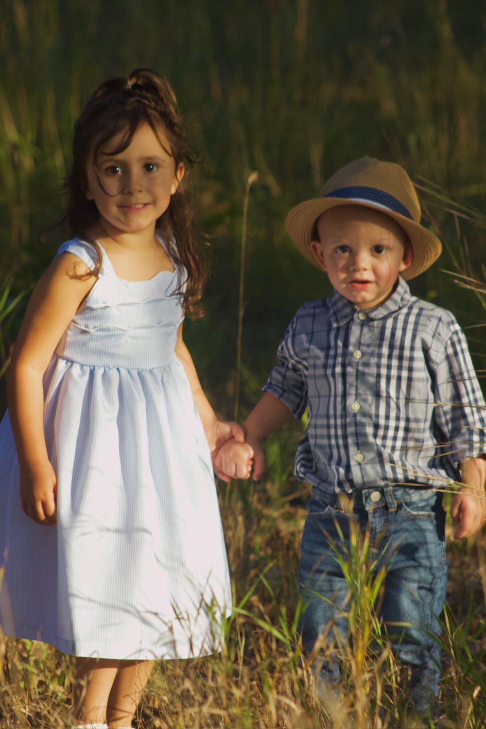Two young children in casual attire pose together outdoors among tall grass at sunset.