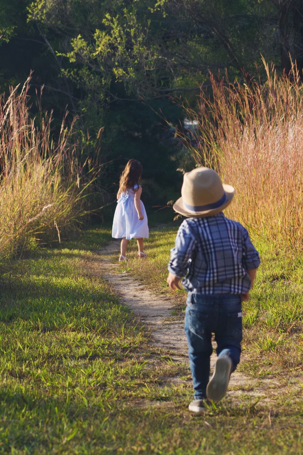 Two small children walk together along a winding dirt path through tall golden grass at sunset.