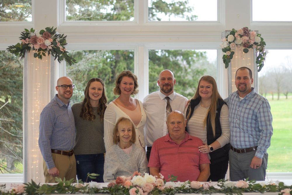 A large group sits together at a long dining table on an outdoor patio at an engagement party for family photos.