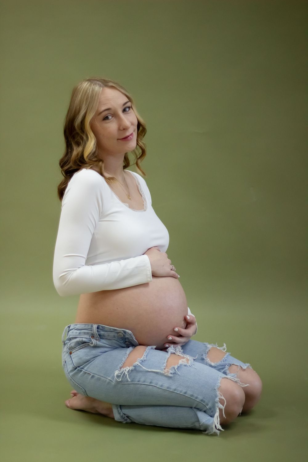 Maternity photos of a woman in a white top and jeans sitting on the floor in front of a green backdrop.