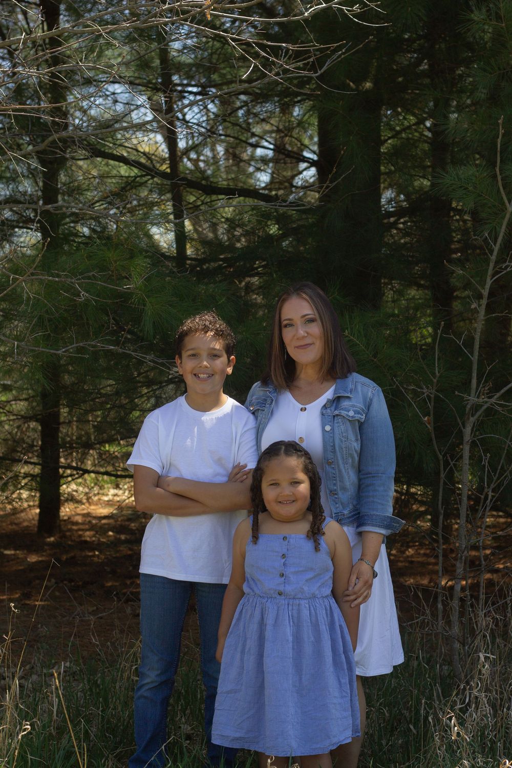 A family poses together in a wooded forest setting on a sunny day, surrounded by tall evergreen trees.