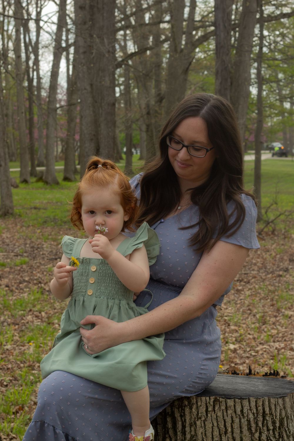 A mother and toddler in a green dress share a snack while sitting on a fallen tree trunk in a wooded area.