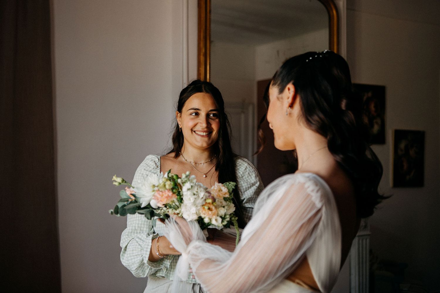 Smiling bride holding a white and peach bouquet while admiring her reflection in an ornate gold mirror.