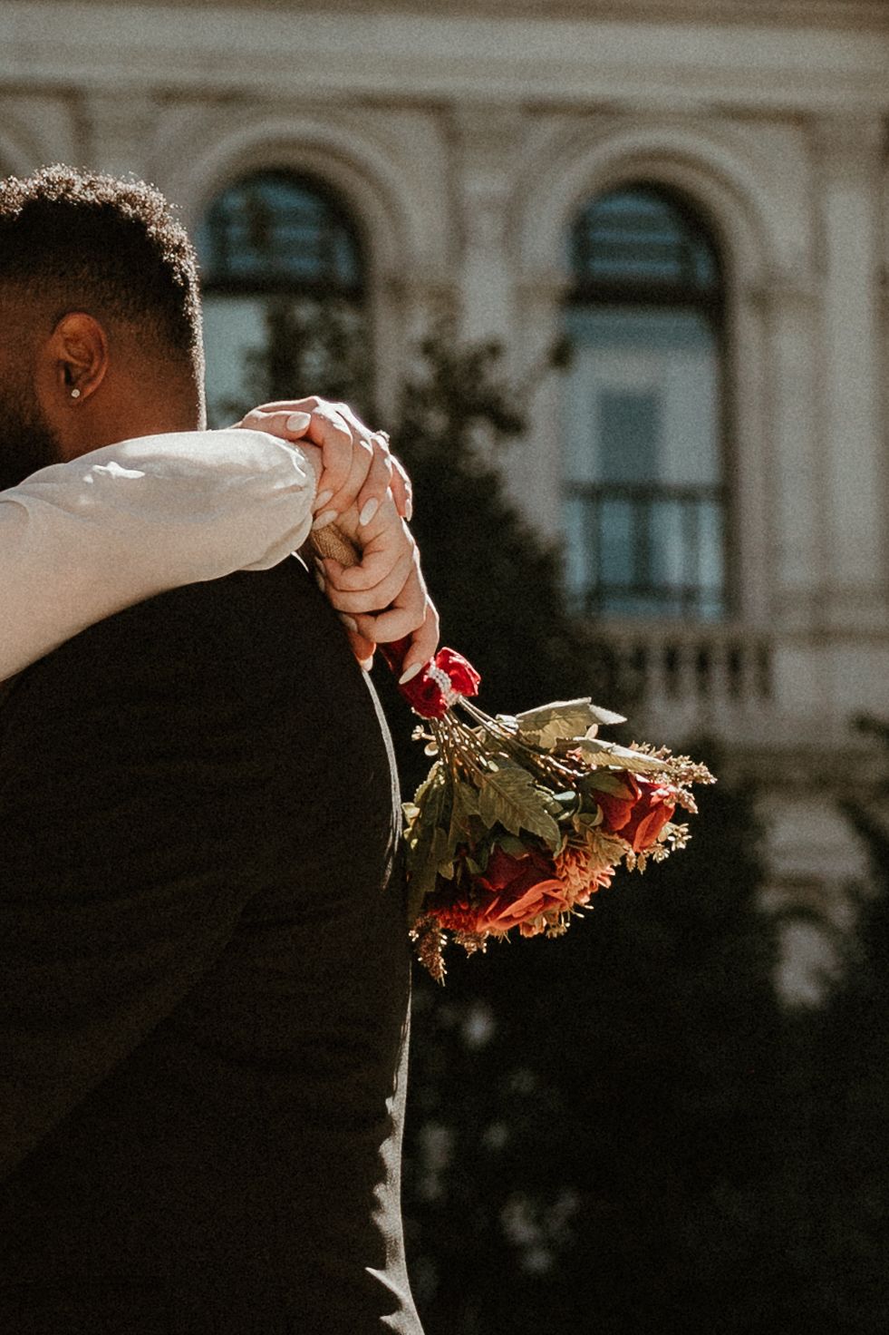 A couple embraces during their wedding photoshoot with a bouquet of flowers against an ornate building facade.