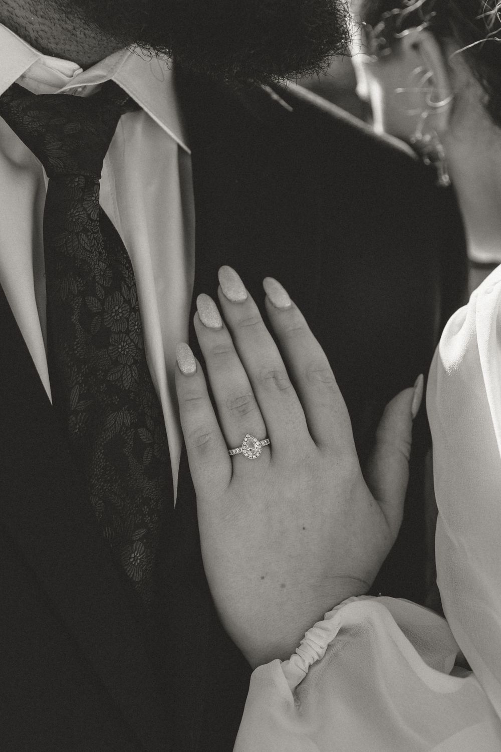 Close-up black and white photo of an engagement ring on a bride's hand against formal attire.