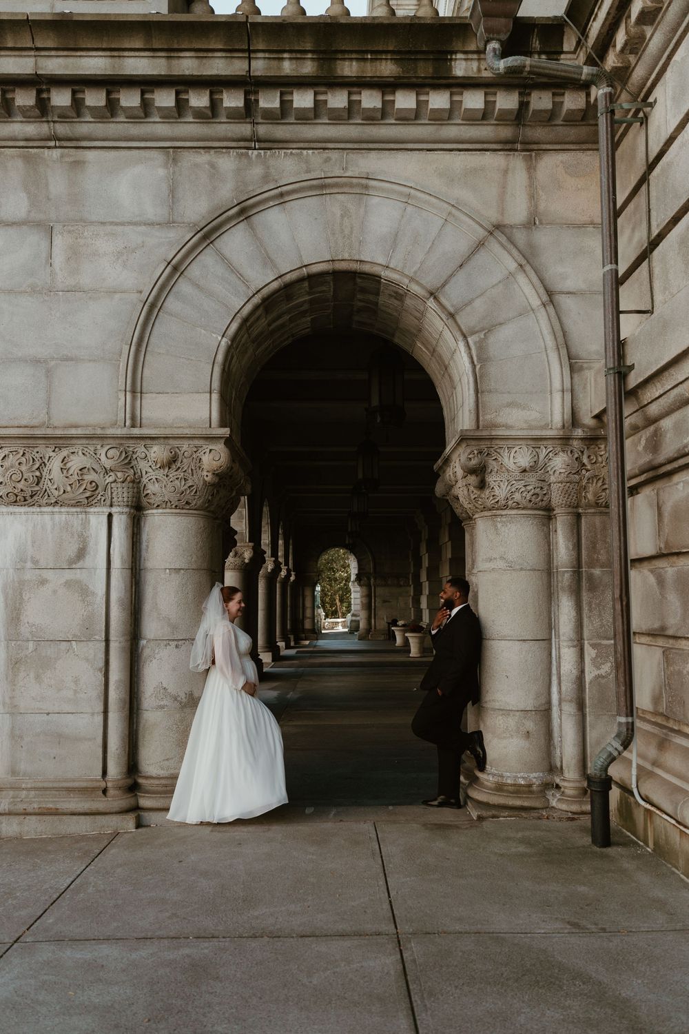 Wedding couple poses in an elegant stone archway corridor with classical architecture.