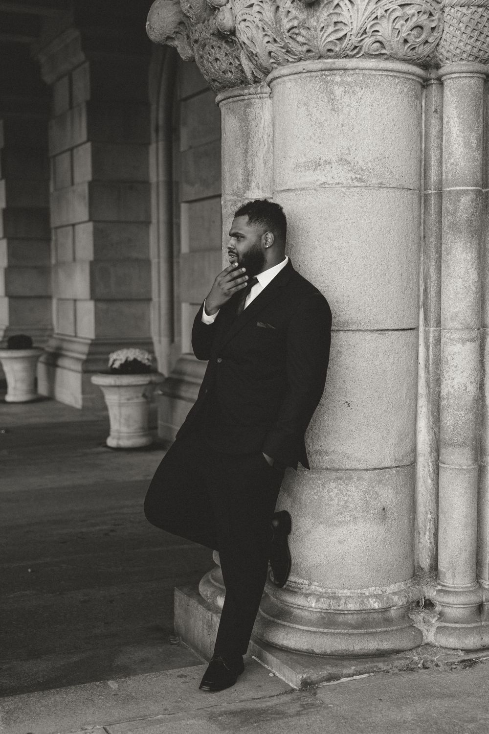 Black and white portrait of a groom in formal attire leaning against an ornate stone column.