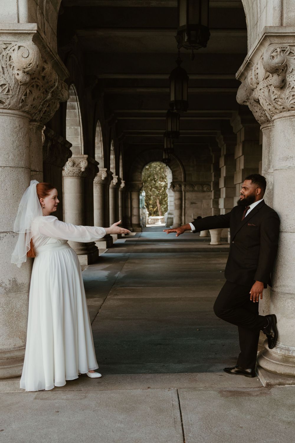 Bride and groom pose playfully in a columned corridor of a historic building.