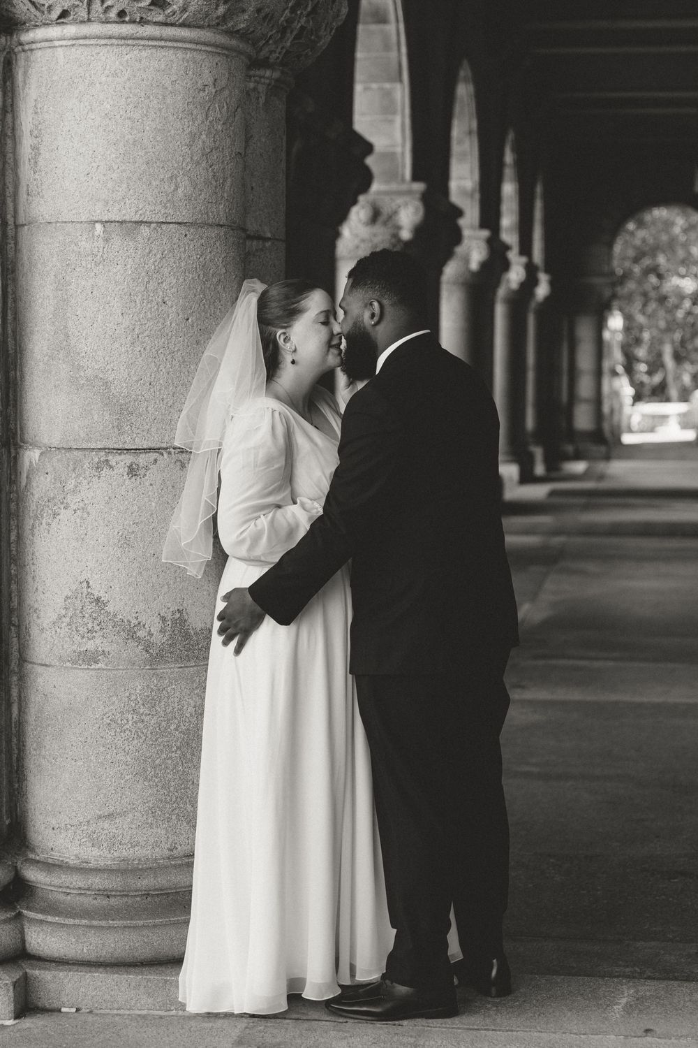 Black and white photo of newlyweds sharing an intimate moment between stone columns.