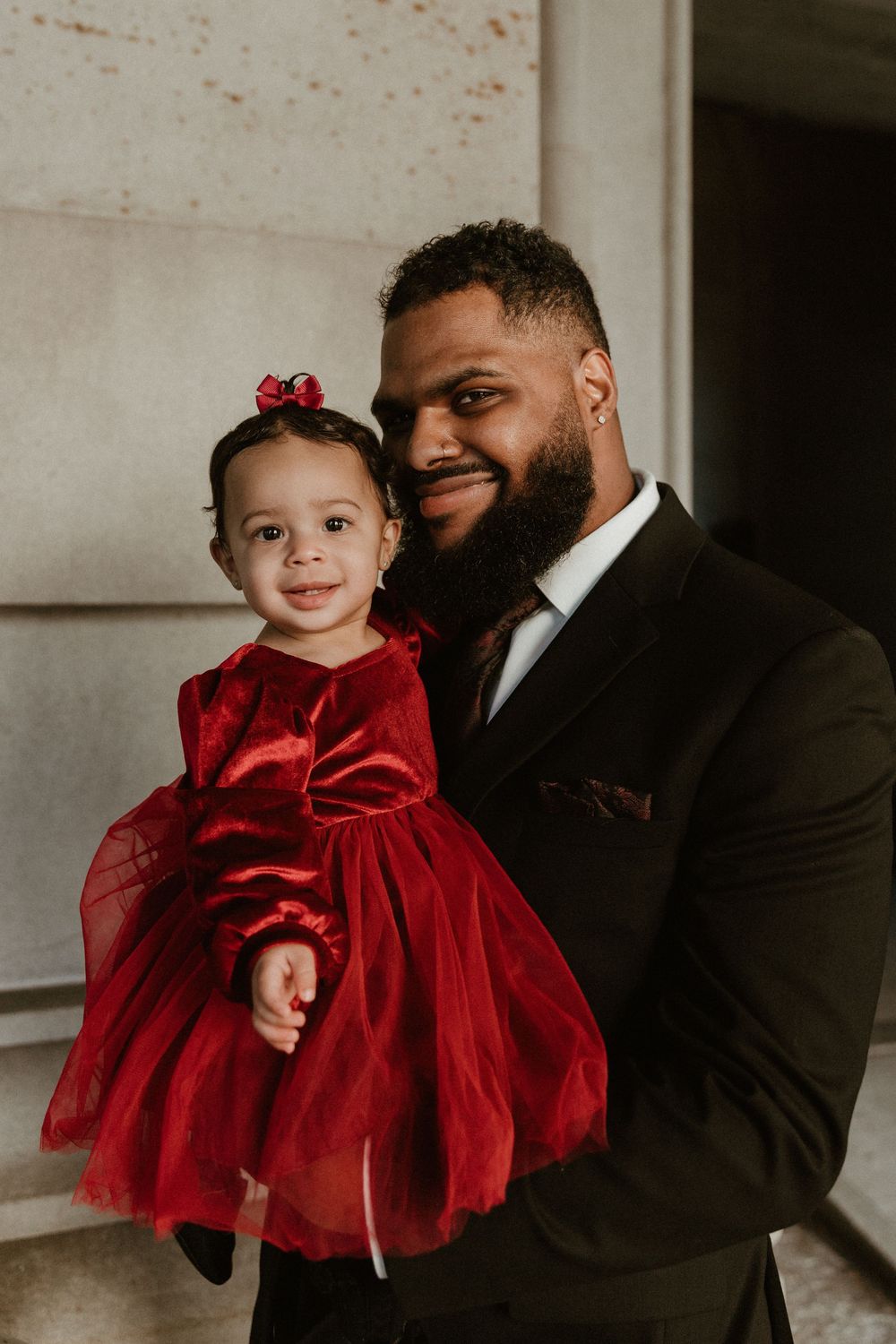 A formal portrait with a little girl in a red dress against a light stone wall.