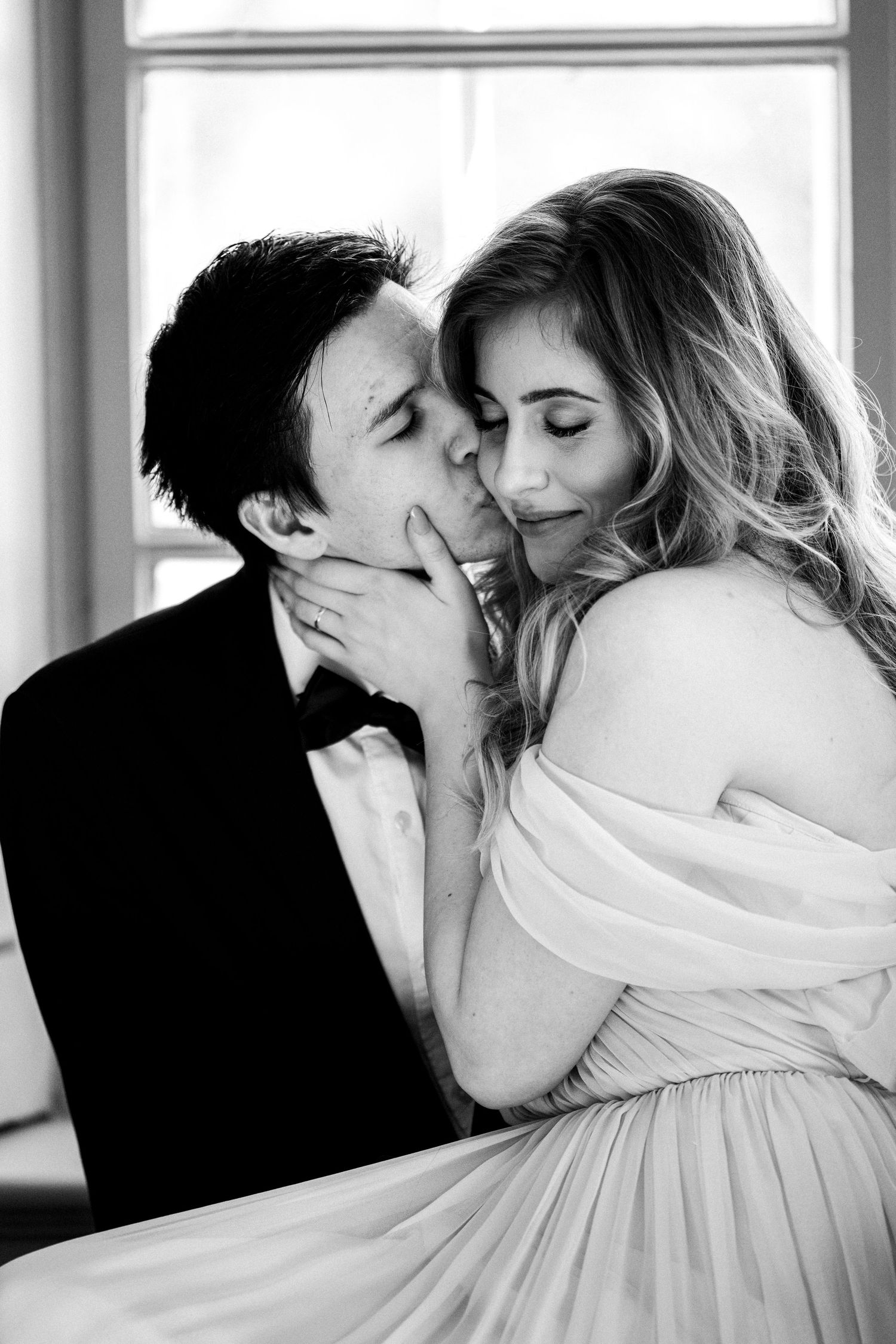A romantic black and white portrait of a couple in formal wear sharing an intimate moment by a window.