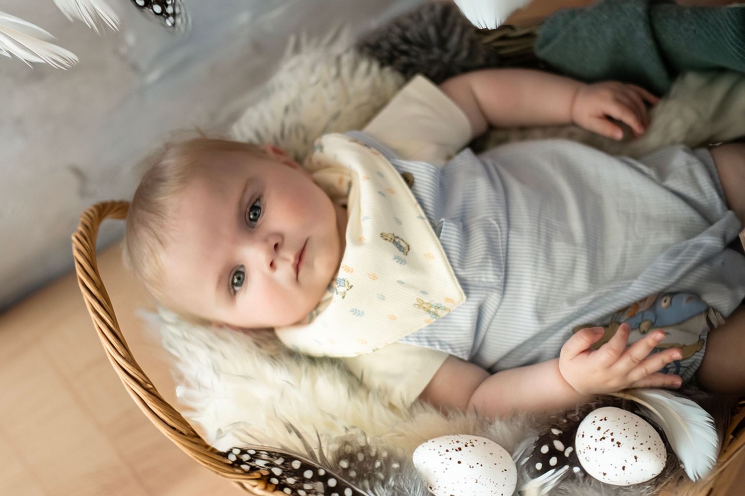 A small baby wearing a white bib rests on a soft beige surface near decorative eggs.