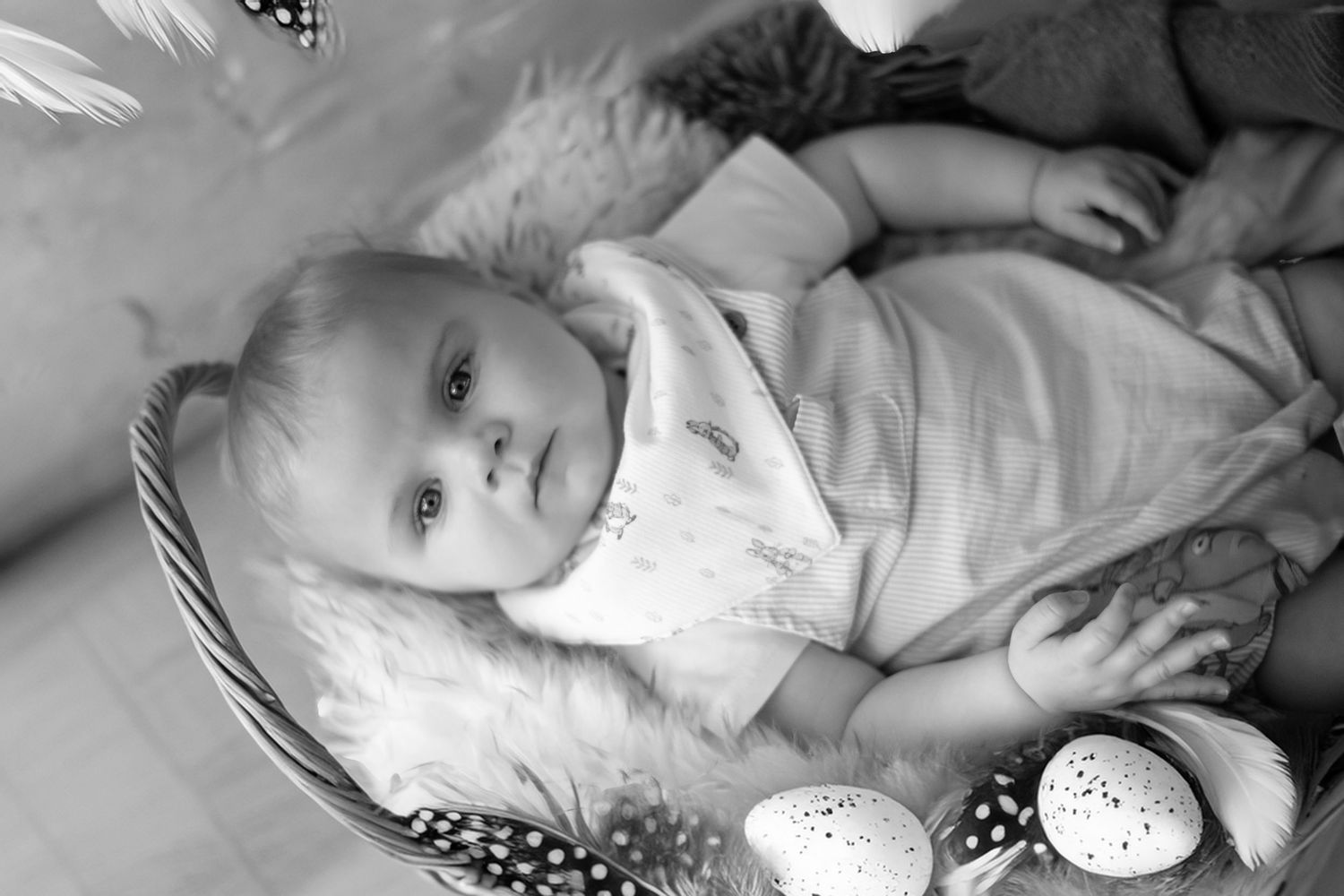 A black and white photograph of a baby lying on a plush surface next to a spotted easter egg.