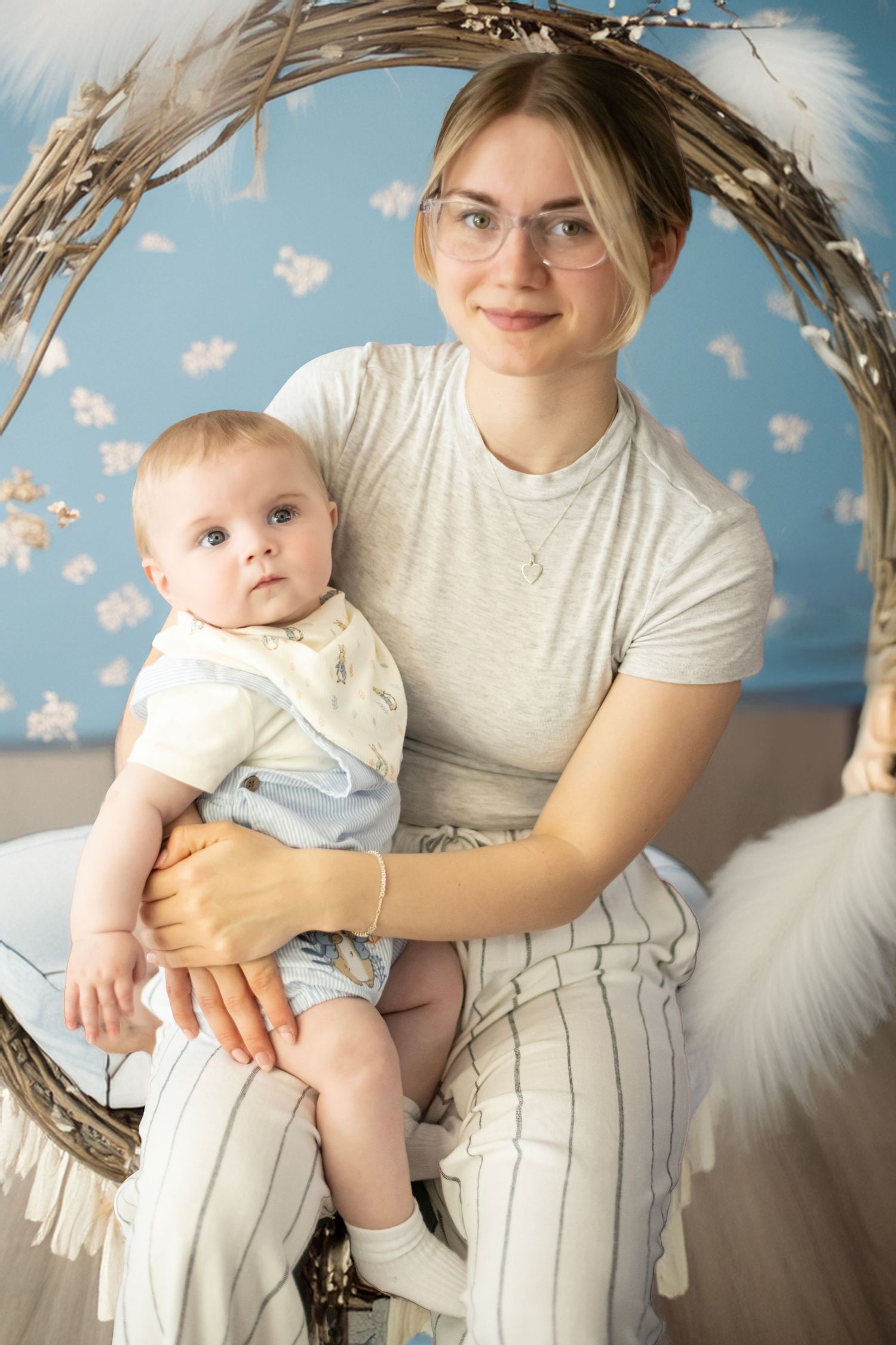 Mother and infant pose together on decorated wicker chair against floral backdrop in soft studio lighting.