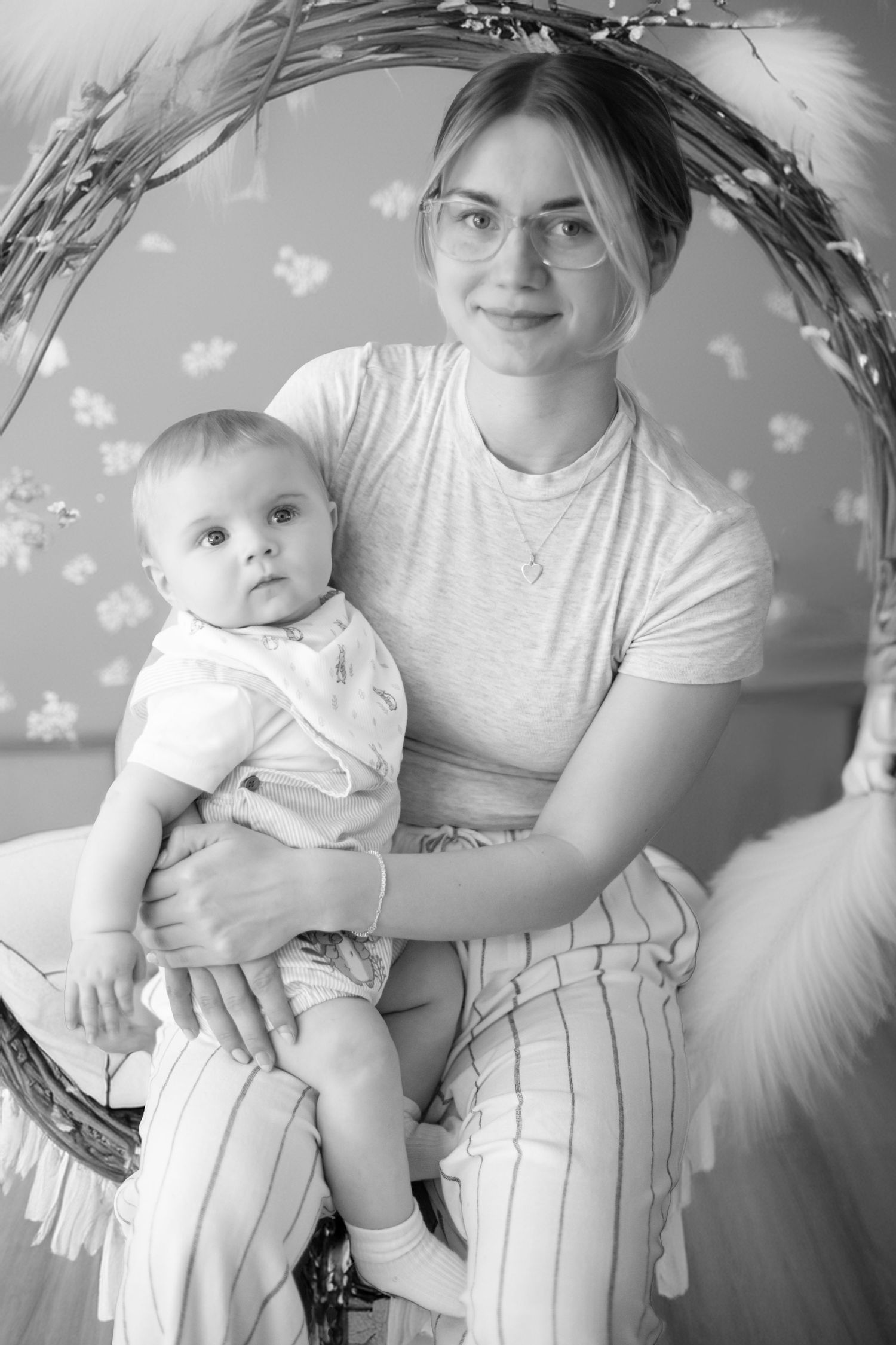 Black and white portrait of parent and baby sitting together in a decorated studio setting with floral patterns.