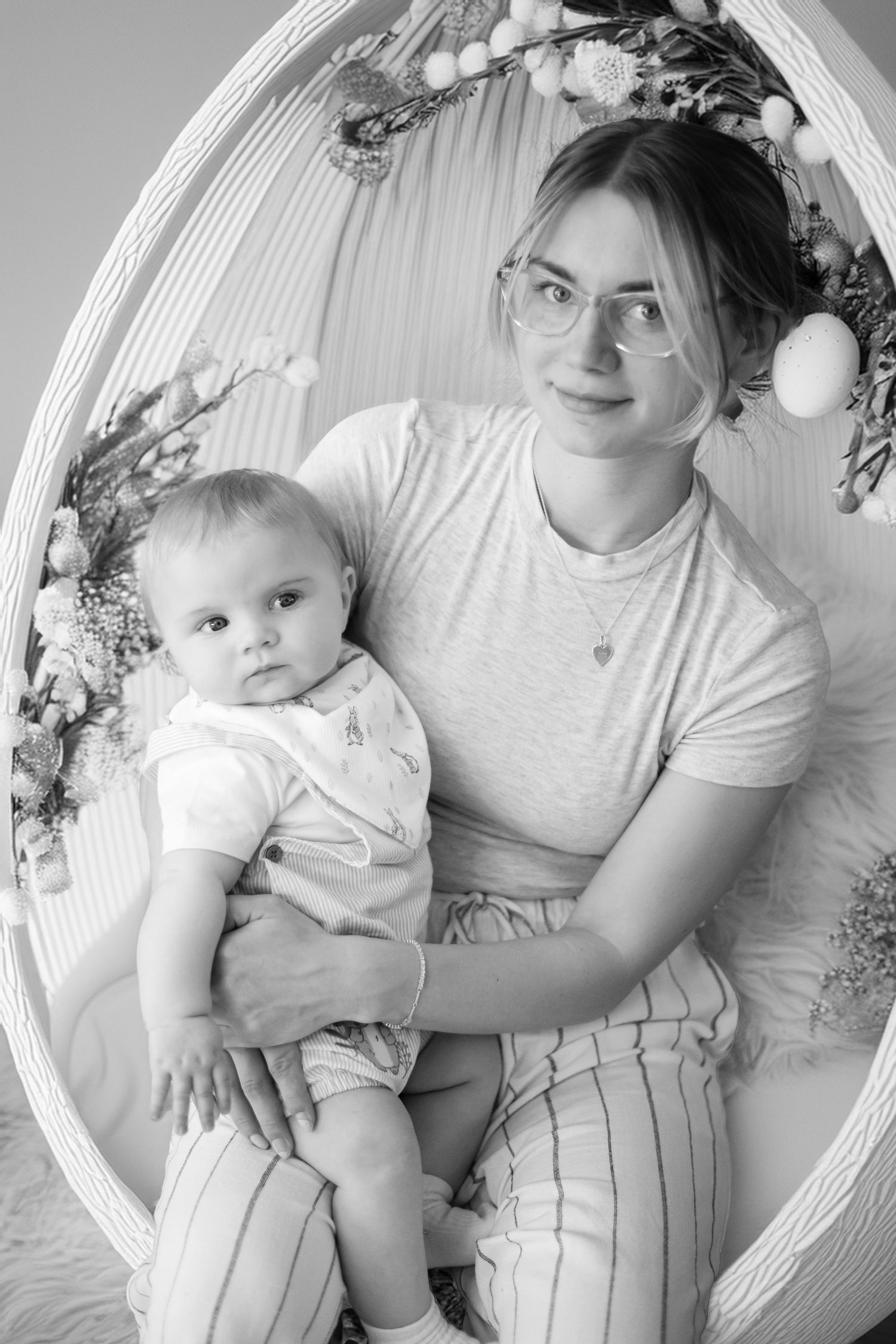 Black and white portrait of mother and baby sitting in ornate wicker chair with floral decorations.