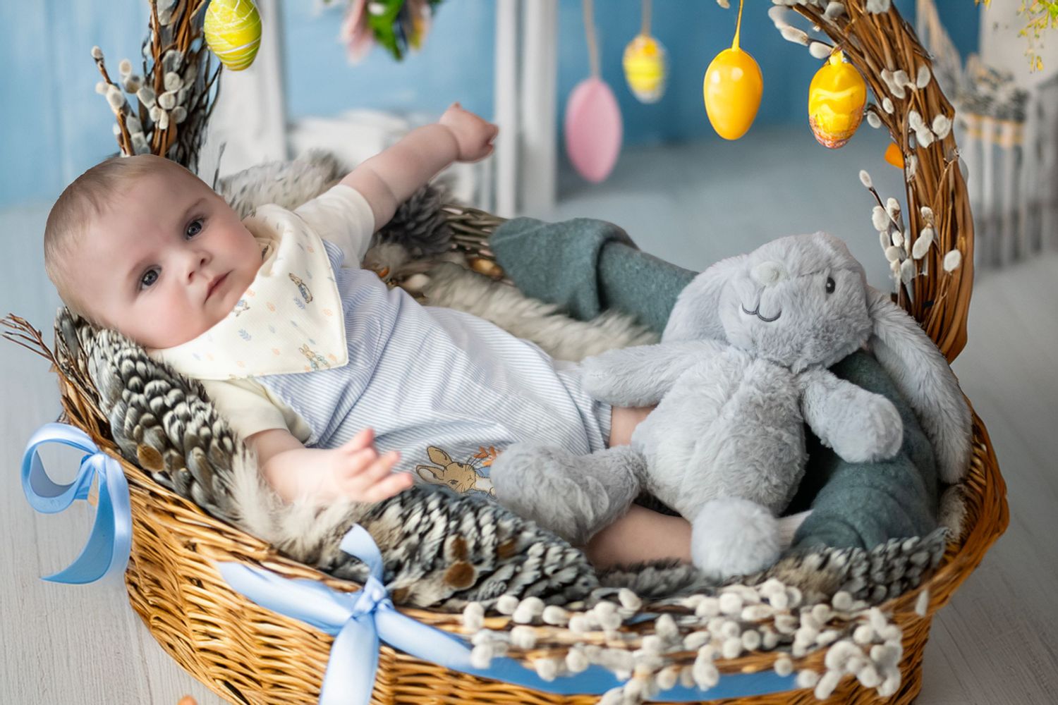 Baby lying in woven Easter basket with stuffed bunny toy and colorful eggs hanging above as decoration.