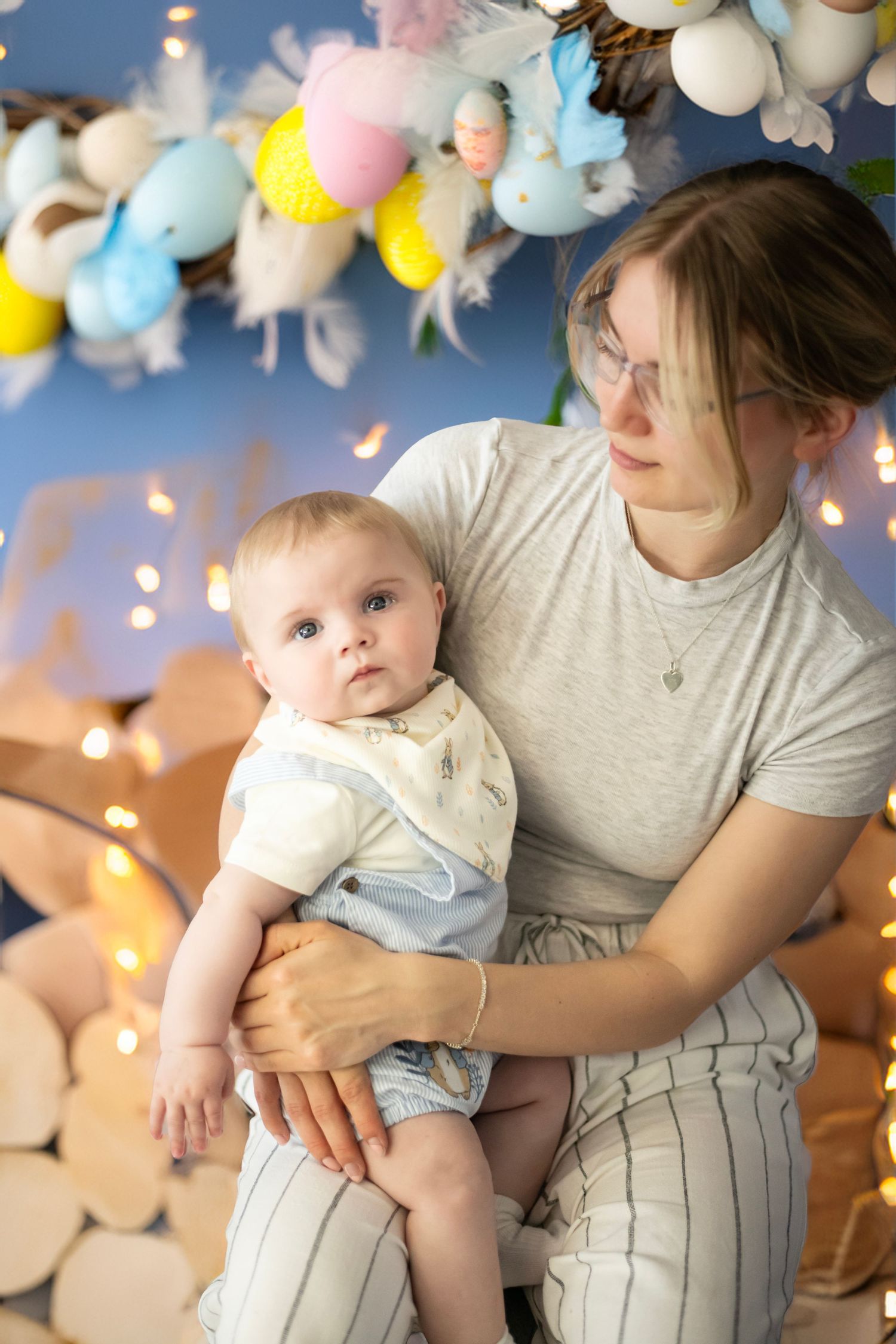 A parent and baby sit together under festive balloon decorations and twinkle lights in a cozy indoor setting.