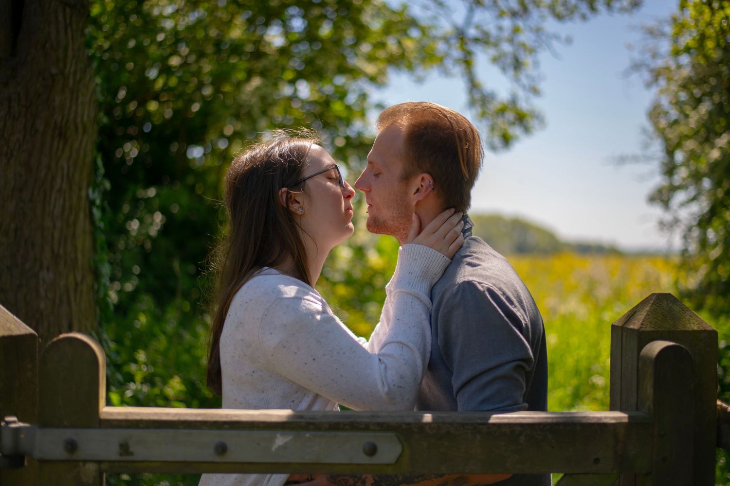 A romantic couple shares an intimate moment on a wooden bench surrounded by sunlit trees and nature.
